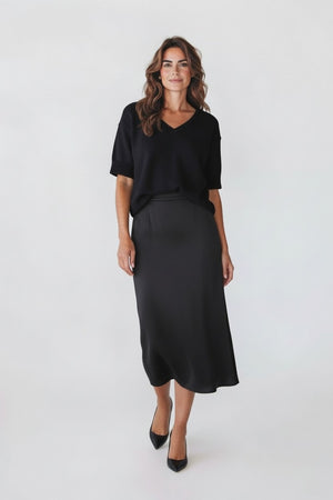A woman with wavy brown hair wears a black short-sleeve V-neck top, black heels, and the ROSE & CROWN ROSE AND CROWN Satin Midi Skirt, standing and smiling against a plain white background.