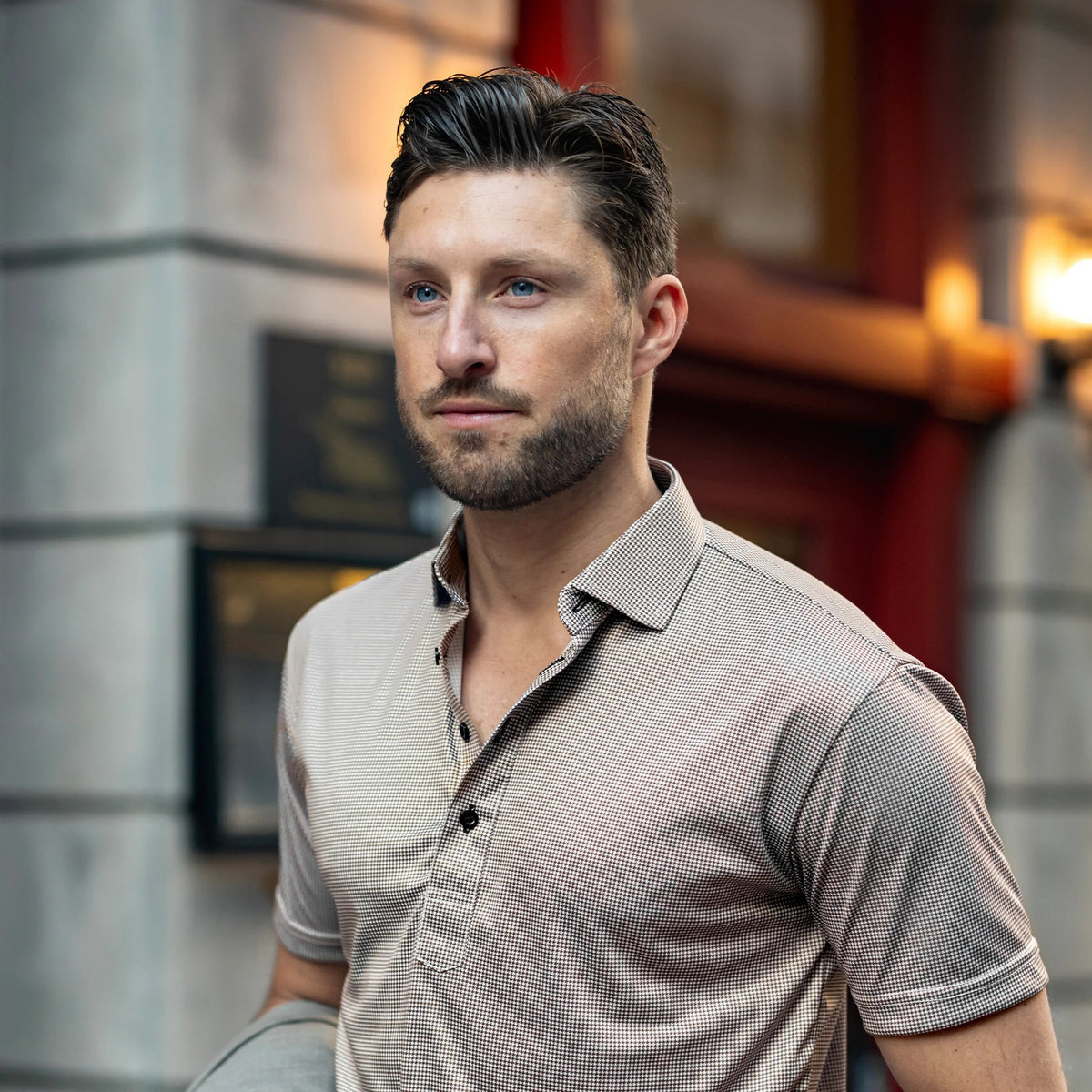 A man with short brown hair and a beard wears the COLLARS AND CO Semi Spread Collar Original Polo in Brown Houndstooth, standing in an urban setting with blurred buildings and warm outdoor lighting in the background.