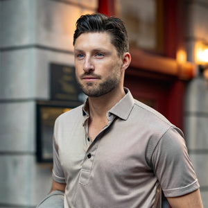A man with short brown hair and a beard wears the COLLARS AND CO Semi Spread Collar Original Polo in Brown Houndstooth, standing in an urban setting with blurred buildings and warm outdoor lighting in the background.