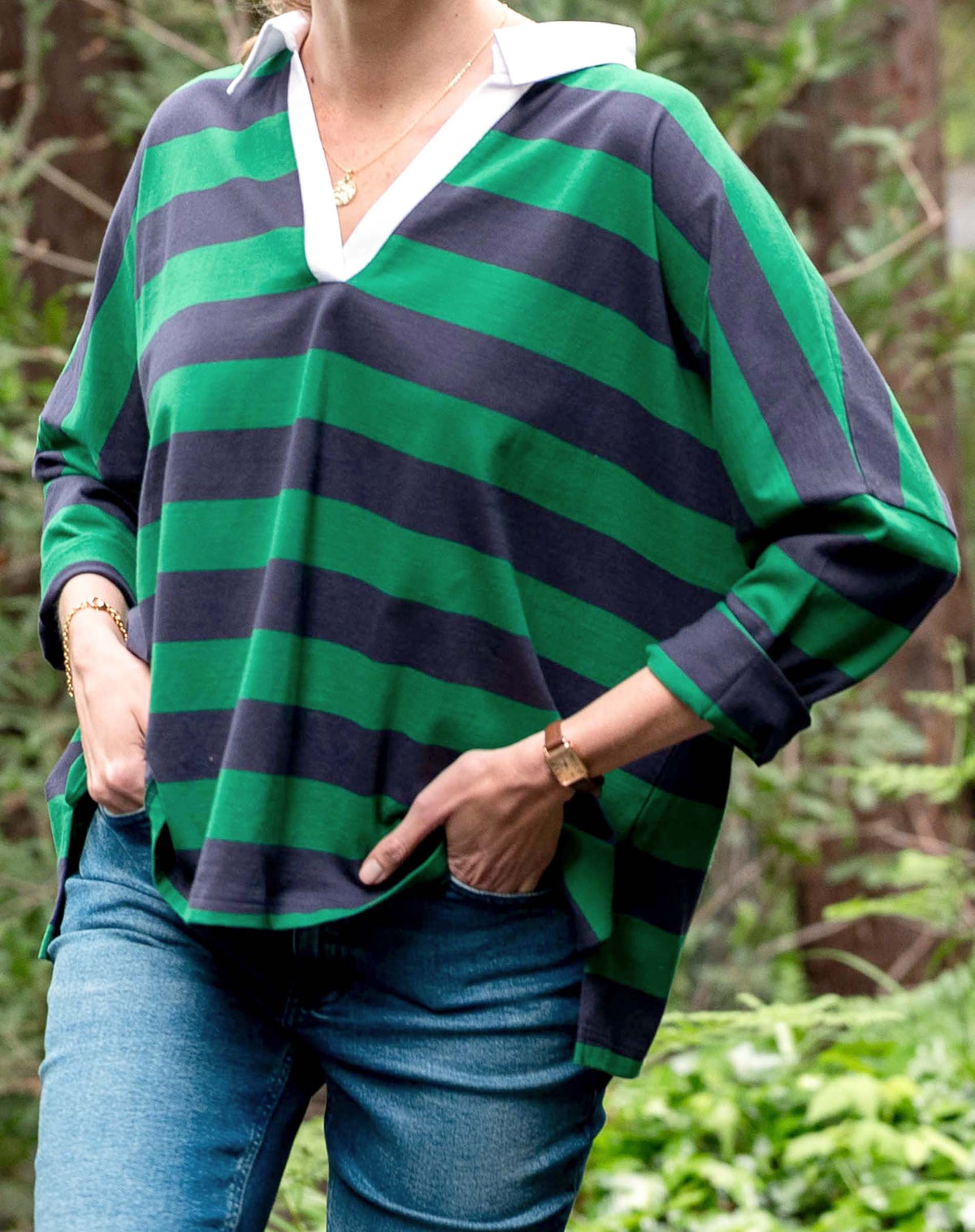 A person wears the MERSEA Amelia Rugby Tee in navy green stripe, featuring a white collar, and blue jeans. They stand outdoors with hands in pockets, green foliage visible behind them.