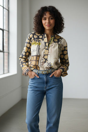 A person with curly hair stands indoors by a window, smiling softly with hands in pockets, dressed in blue jeans and the STORIATIPIC - MARTGOT BLOUSE IN DELTA by STORIATIPIC—a patterned cotton button-up featuring abstract designs. This blouse is easy to care for: simply machine wash gently at 30 degrees to keep it looking fresh.