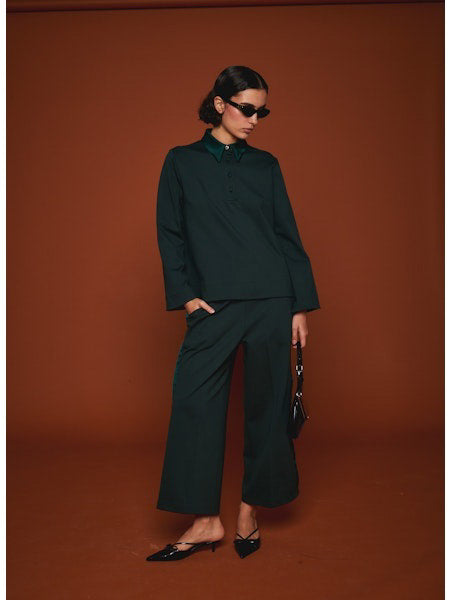 Facing away from the camera, a person wears MILVA’s MILVA MI - TUXEDO DRESS PANT in dark green with cropped legs and black heels, holding a black handbag. The background is solid brown.
