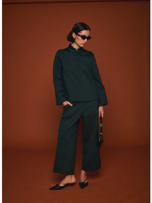 Facing away from the camera, a person wears MILVA’s MILVA MI - TUXEDO DRESS PANT in dark green with cropped legs and black heels, holding a black handbag. The background is solid brown.