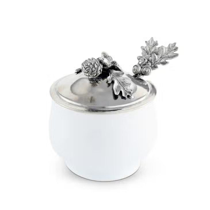 The ACORN OAK LEAF SUGAR BOWL by VAGABOND HOUSE is a small fine porcelain bowl with a silver lid adorned with intricate decorative elements, including acorns and oak leaves. The lid&#39;s handle is an ornate oak leaf design, enhancing the bowl&#39;s elegant and detailed appearance, reminiscent of a luxury grade pewter Antler Creamer Set.