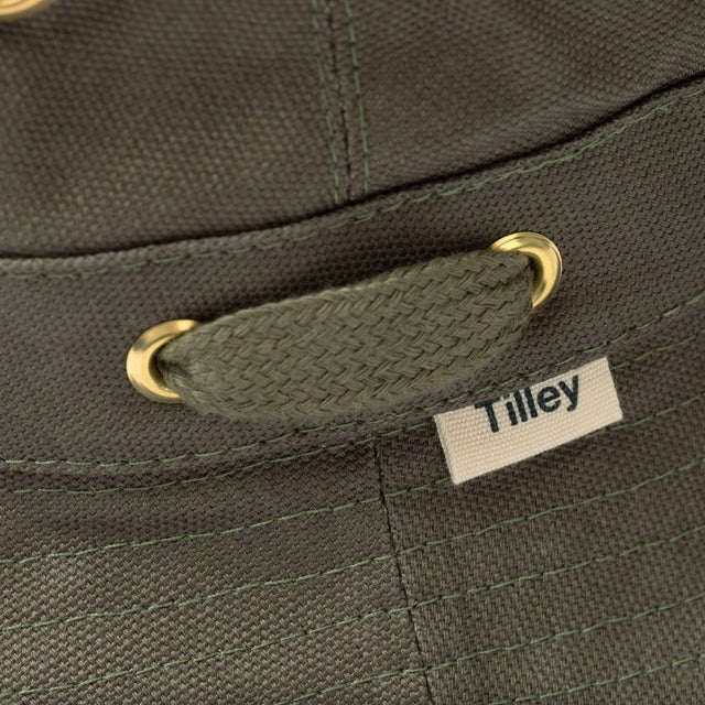 A person wearing the TILLEY - ICONIC T1 BUCKET HAT IN OLIVE with yellow eyelets and a white crew-neck shirt looks slightly to the side against a plain light background. The TILLEY hat offers UPF 50+ sun protection.