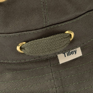 A person wearing the TILLEY - ICONIC T1 BUCKET HAT IN OLIVE with yellow eyelets and a white crew-neck shirt looks slightly to the side against a plain light background. The TILLEY hat offers UPF 50+ sun protection.