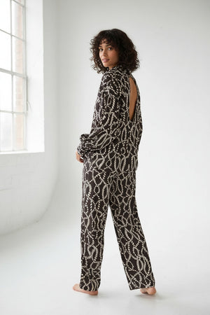 A woman stands against a light background wearing the SEVENTY VENEZIA Printed Shirt with Open Back by SEVENTY, styled with matching black and white polka dot pants, layered necklaces, and open-toed shoes. With her hands in her pockets and a relaxed expression, she showcases the shirt’s lightweight fabric, thoughtfully crafted for both comfort and style.