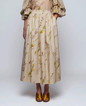 A woman stands against a plain background wearing the MIRTO 1956 Beige Floral Print Midi Skirt, paired with puffed three-quarter sleeves, a wide tan belt, and brown open-toe heels. Her hair is styled in a high bun with a decorative accessory.