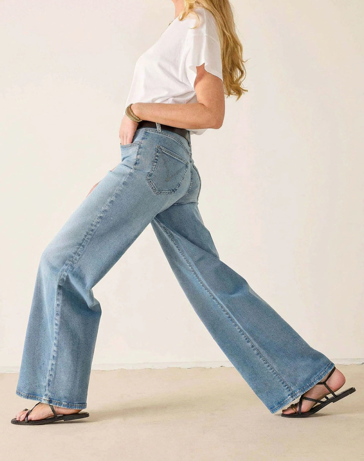 A woman stands against a plain background wearing a white T-shirt, light blue MERSEA KATE WIDE LEG JEANS IN CLOUD NINE featuring four-way stretch and tummy tuck technology, a black belt, black sandals, and a gold necklace, with her hands resting on her hips.