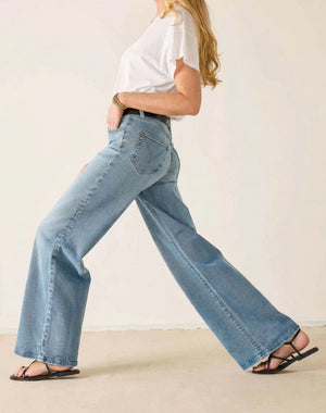 A woman stands against a plain background wearing a white T-shirt, light blue MERSEA KATE WIDE LEG JEANS IN CLOUD NINE featuring four-way stretch and tummy tuck technology, a black belt, black sandals, and a gold necklace, with her hands resting on her hips.