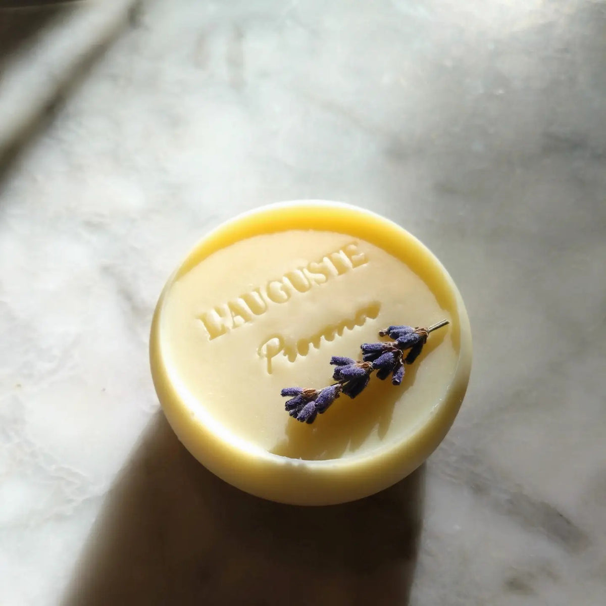 L&#39;AUGUSTE PROVENCE - THE ESSENCE OF PROVENCE LAVENDER SOAP is a round, handmade French soap wrapped in painted landscape fabric, presented in a Provence gift box. Dried lavender rests nearby on a white cloth background.