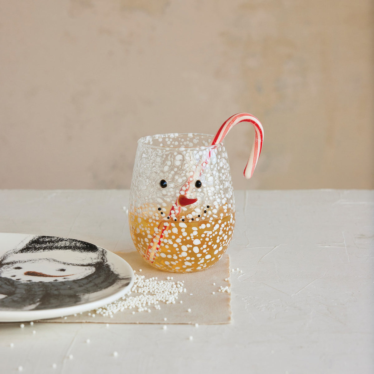 The CREATIVE COOP CONFETTI DRINKING GLASS WITH SNOWMAN FACE is a clear, stemless glass with a textured, bubbly surface and a cheerful snowman face featuring black dot eyes and mouth plus an orange-red curved nose.