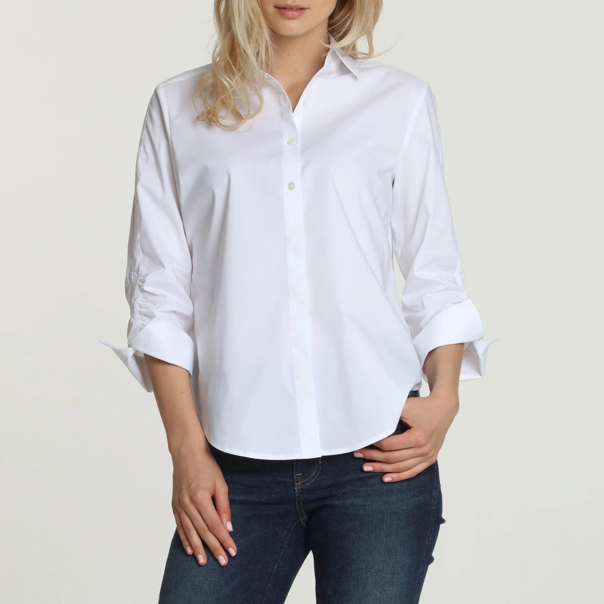 A woman wearing the HINSON WU - ZOEY 3/4 SLEEVE SHIRT WITH RUCHED SLEEVE by HINSON WU, a refined white button-up shirt made from a premium cotton blend with 3/4 ruched sleeves, paired with dark blue jeans. She stands against a plain light background with her hands resting near her hips, showcasing the shirt’s stylish look and practical machine-washable design.