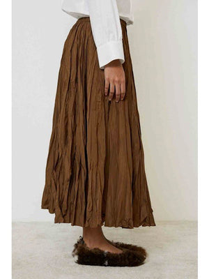 A person wearing the MEIMEIJ CRINKLE SKIRT stands against a plain light background. Only the lower half, featuring the brown midi skirt and brown fuzzy slippers, is visible.