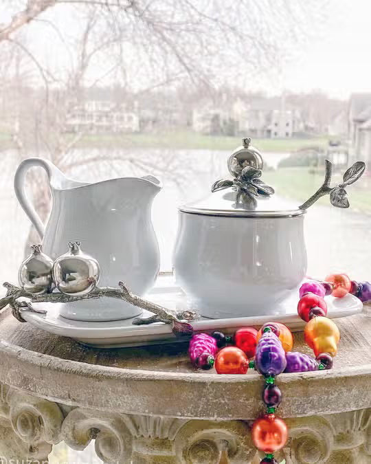 The VAGABOND HOUSE POMEGRANITE CREAMER SET includes a white ceramic sugar bowl with a metallic lid featuring pewter pomegranates, a matching cream pitcher, and a silver spoon with branch-like handle and pomegranate accents on a rectangular tray.
