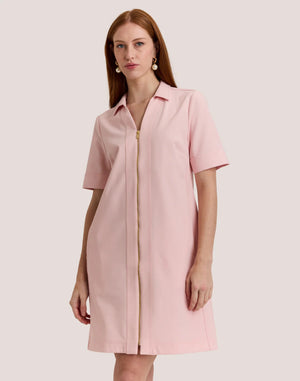 A woman with long, straight auburn hair wears the HINSON WU Bridget Short Sleeve Traveler's Twill Dress in blush pink, featuring a collar and gold front zipper, standing against a plain light background.