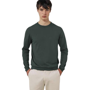 A young man stands with his hands in his pockets against a white background, wearing the HARTFORD Extra-Fine Merino Wool Crew Pullover in Forest and light-colored pants.