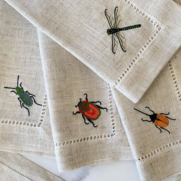 The DOT AND ARMY - EMBROIDERED INSECT DINNER NAPKIN by DOT AND ARMY features a beige linen blend material and showcases embroidered insects—including a green beetle, red beetle, orange beetle, and a dragonfly—overlapping on a white surface. These reusable napkins bring a whimsical touch to any table setting.