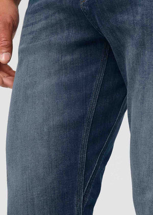 A person wearing a white t-shirt, DUER PERFORMANCE DENIM ATHLETIC TAPER PANTS IN GALATIC, and white sneakers stands facing away from the camera against a plain light background.