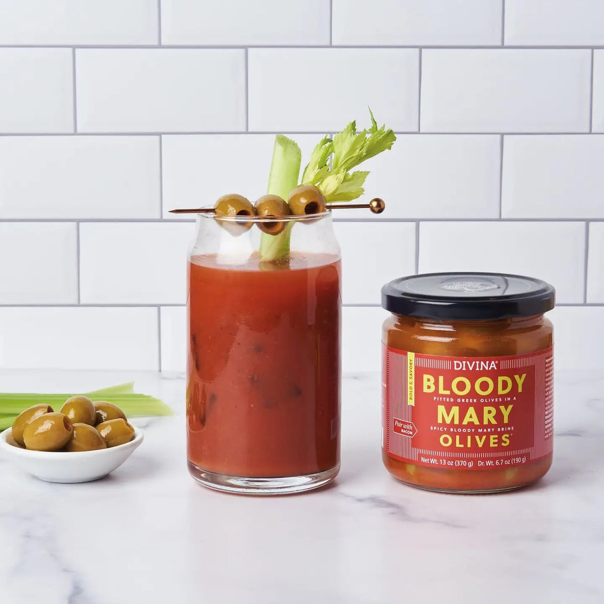 A Bloody Mary cocktail garnished with celery and DIVINA - BLOODY MARY OLIVES sits next to a jar of DIVINA - BLOODY MARY OLIVES and a small bowl of green olives on a white countertop with a tiled background, highlighting the perfect cocktail garnish enhanced by flavorful Bloody Mary brine from DIVINA.