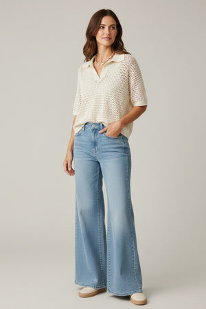 A woman stands against a plain background, wearing the BASE MILANO Short Sleeve Splitneck Cotton Polo in Ivory with light blue high-waisted wide-leg jeans, beige shoes, and a necklace. She has brown wavy hair and poses with one hand in her pocket for a chic BASE MILANO-inspired look.