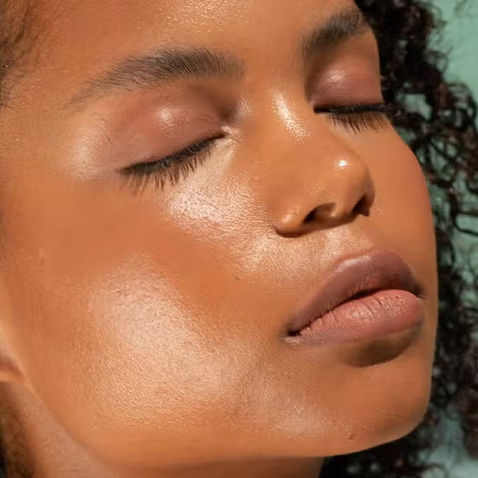 A close-up of a person with curly dark hair, a serious expression, and warm makeup featuring the universally flattering, weightless JASON WU - HOT FLUFF LIP/EYE/CHEEK by JASON WU BEAUTY on their face. They are looking directly at the camera with a neutral background.