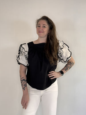 A person with long brown hair and tattoos on both arms stands against a plain background, smiling. They wear the FARYLINA - DAHLIA TOP by FARYLINA—a black and white statement blouse with ruffled sleeves—white pants, and an Apple Watch.