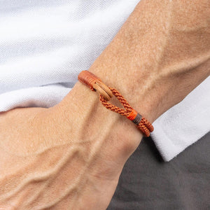 The MAKARLA MEAN'S NAUTICAL ROPE BRACELET IN CHOCOLATE, handmade and adjustable, is displayed on a white cylindrical stand with a label beneath it matching the bracelet’s color.
