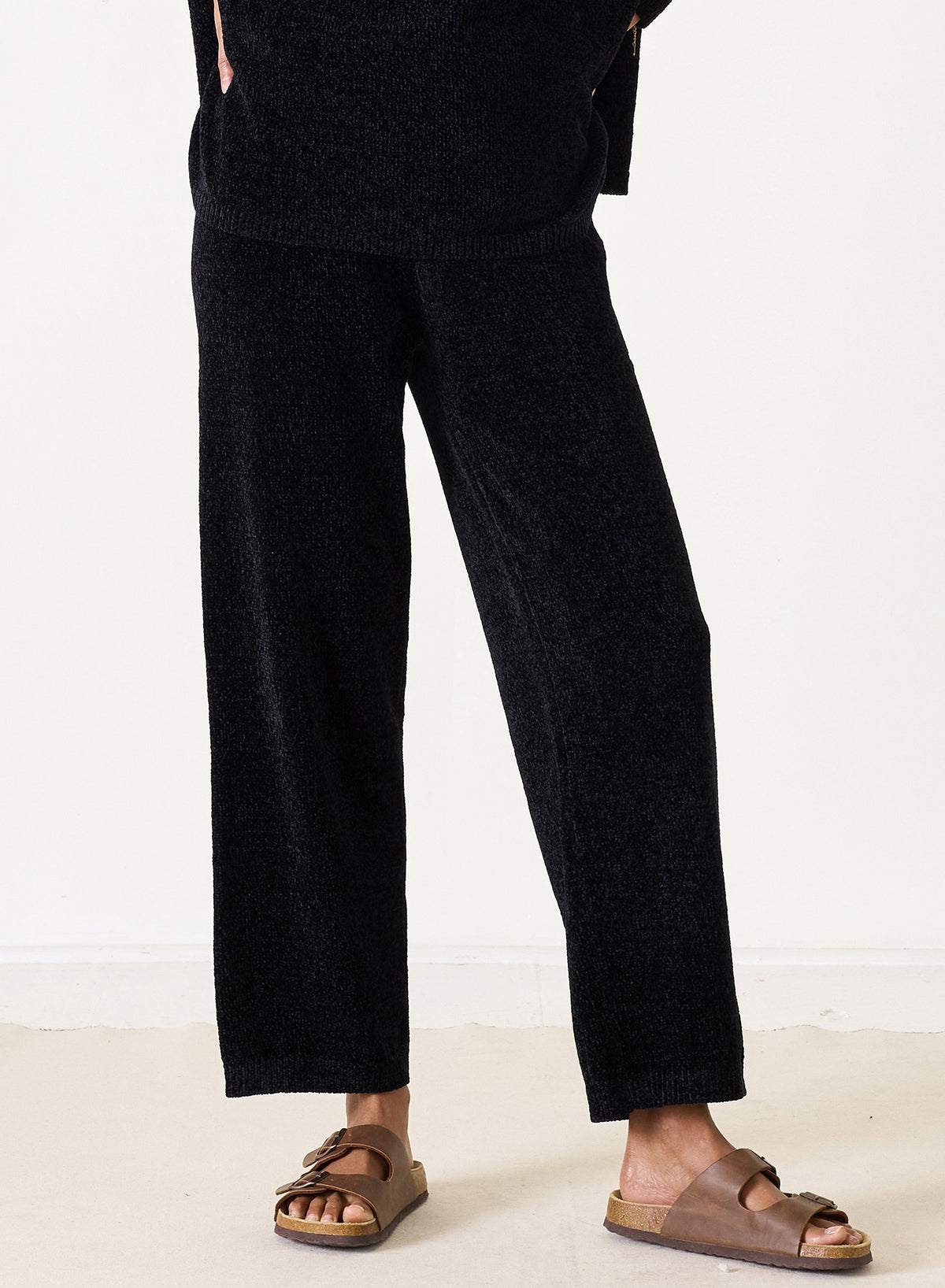 Someone stands indoors on a light floor against a white wall, wearing the MERSEA - EVERSOFT CHENILLE PANT in black with a matching top and brown slide sandals.