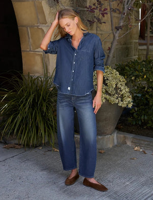 A close-up of a person wearing the FRANK & EILEEN Brennan Everyday Barrel Jean in California Vintage Denim 1993 Wash, paired with a loose blue shirt and one arm resting by their side.