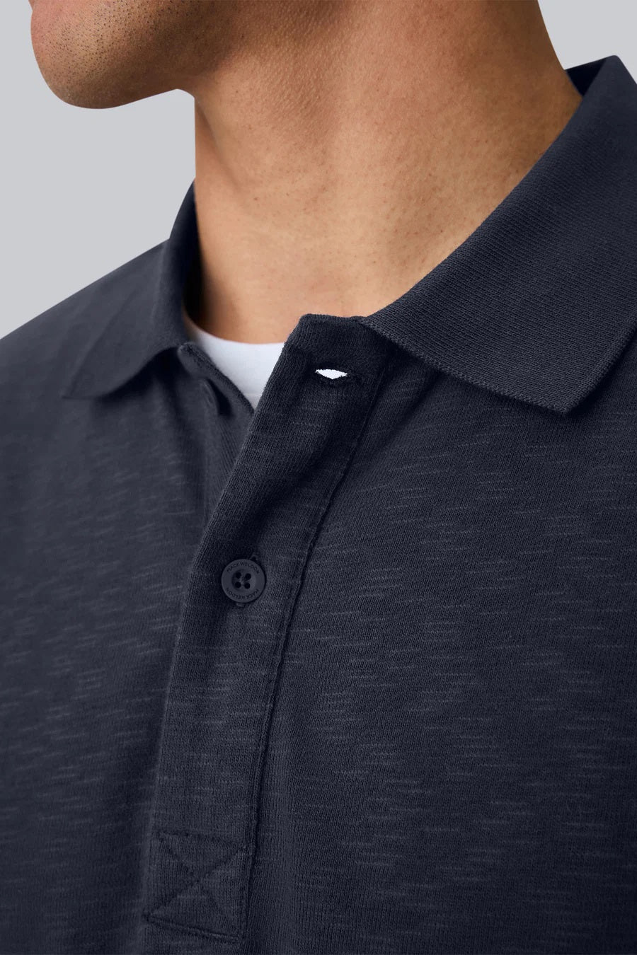 A man with short dark hair is seen from behind, wearing the MACK WELDON - HEAVYWEIGHT SLUB LONG SLEEVE POLO IN FROTH and blue pants, standing against a plain light gray background.