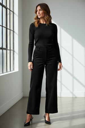 A woman poses against a plain background wearing the SPANX - CORDUROY WIDE LEG JEAN IN BLACK, paired with a fitted black long-sleeve top, black belt, and black heels, her hands in her pockets and expression neutral.