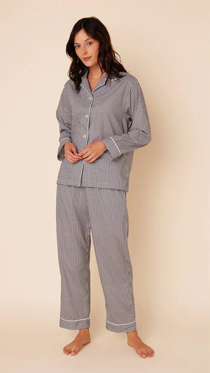 A person with long dark hair is standing on a light beige background, wearing a matching set of loose-fitting, long-sleeved CAT'S PAJAMAS - CLASSIC GINGHAM LUXE PIMA PAJAMA by CAT'S PAJAMAS. Made from Luxe Pima Cotton, the buttoned-up pajamas feature a collar. The person is barefoot and looking slightly towards the camera.