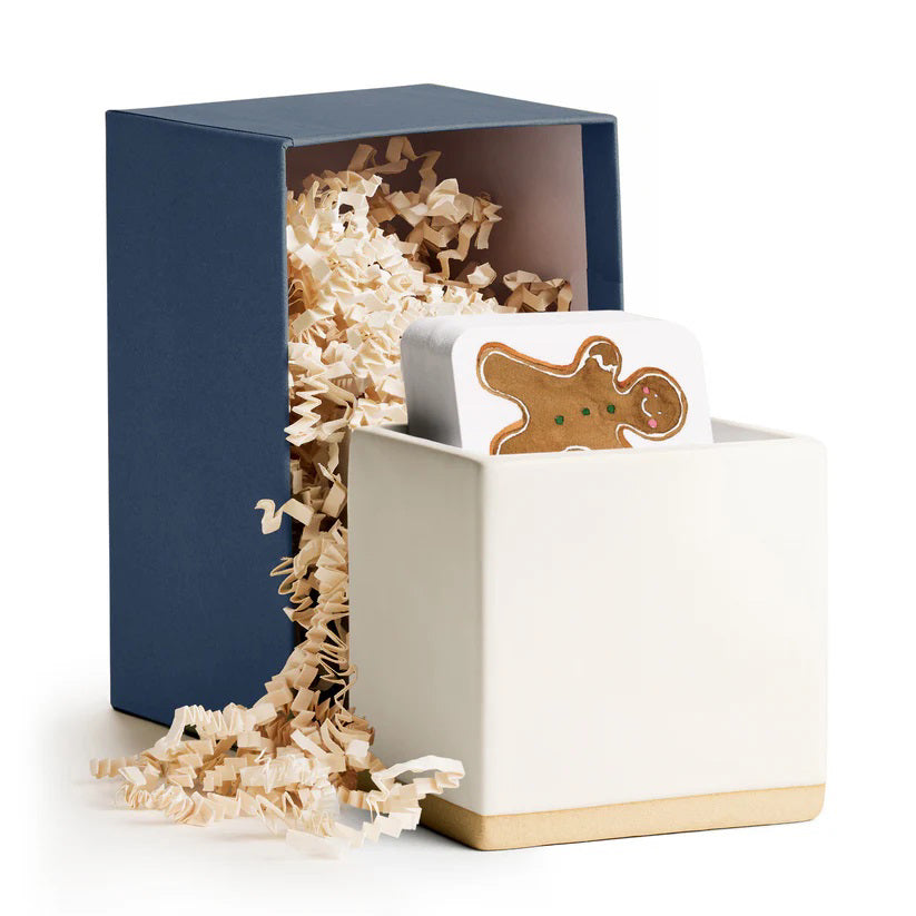 The E. FRANCES PAPER - LITTLE NOTES CERAMIC HOLDER - WHITE, designed as a modern minimalist ceramic cube gift box, contains decorative shredded paper and a gingerbread man-shaped white object, with the partially opened container revealing more shredded paper spilling out.