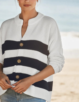 A woman stands on a beach wearing the FRANK & EILEEN Montecito Crew Neck Cardigan in Pure Italian Cotton, featuring navy and white stripes in a relaxed fit with large buttons, paired with light blue jeans. The ocean and sky are softly blurred in the background.