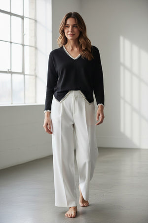 A person with light blonde hair stands sideways against a white background, wearing the MEIMEIJ V Neck Hi Low Top with embroidered contrast stitch detail by MEIMEIJ, paired with white pants.