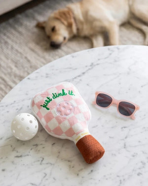 The HAUTE DIGGITY DOG PINK CHECKER PICKLEBALL PADDLE DOG TOY is a plush, paddle-shaped squeaky dog toy featuring a fun pink checkered design, an embroidered pickleball, the phrase "just dink it." in green, and a brown handle—perfect for pups who want to join in on your pickleball passion.