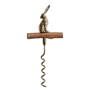 A bottle of wine with a golden foil sits beside the BE HOME - HARE CORKSCREW by BE HOME, INC, featuring a brass rabbit-shaped handle and wooden grip on a light wood surface—an elegant gift for any hostess.