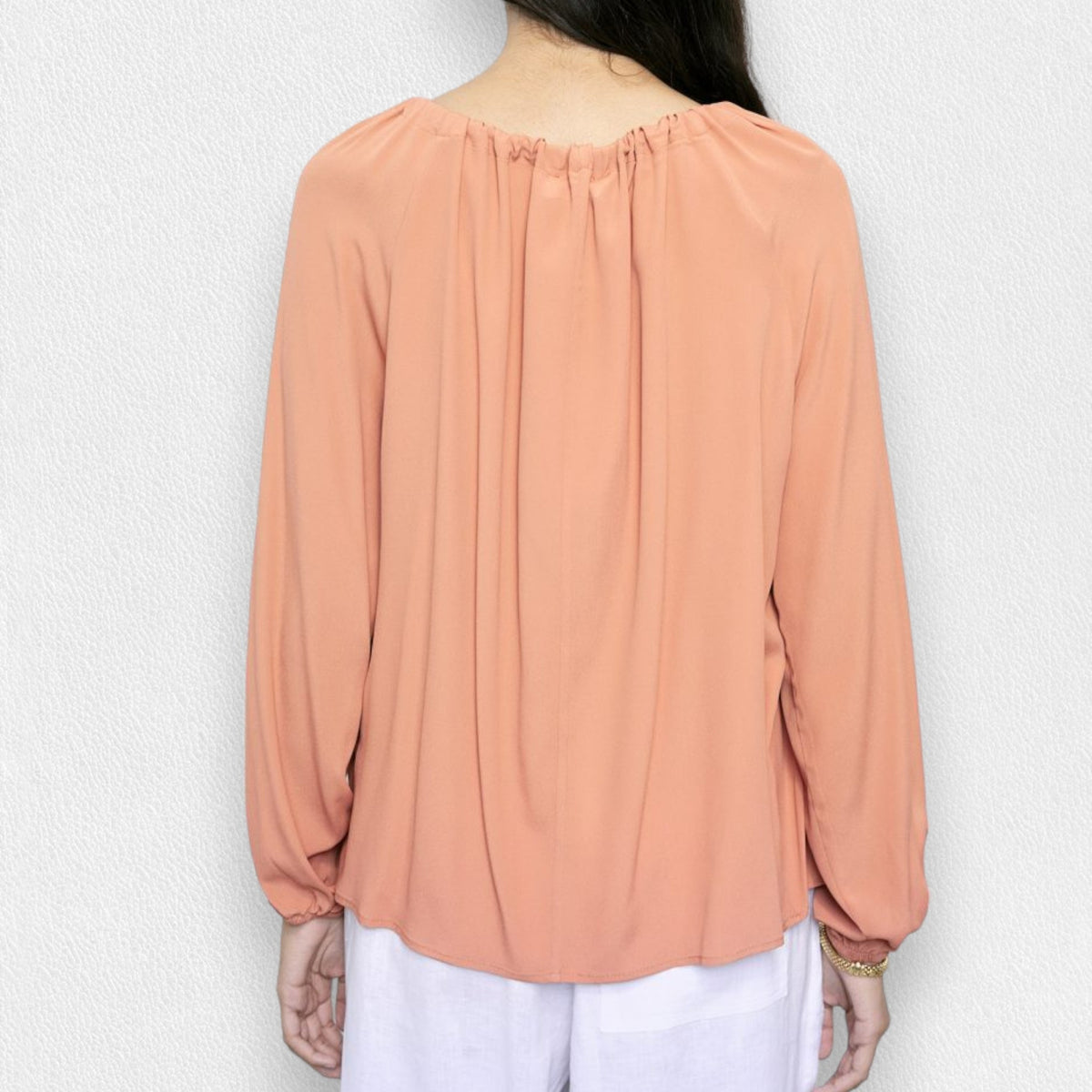A person wearing a MEIMEIJ - TIE NECK BLOUSE in salmon color, paired with white pants, stands against a light gray textured background. The blouse features long sleeves and a bow at the neckline. The person's face is not visible in the image.