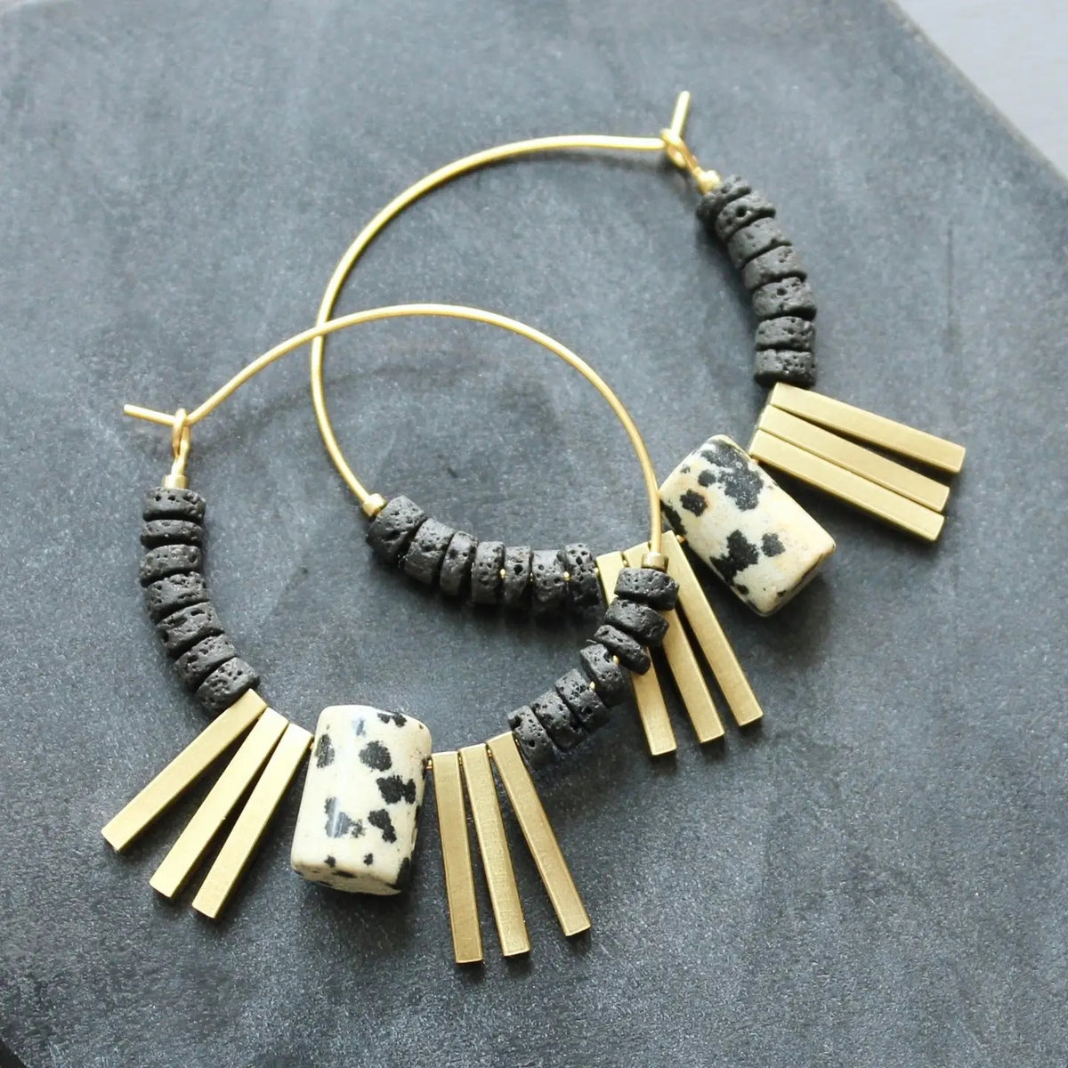 The DAVID AUBREY - Lava Bead and Dalmatian Jasper Hoop Earrings by DAVID AUBREY feature gold plated hoops adorned with black lava bead accents, a rectangular Dalmatian jasper stone, and multiple gold rectangular charms, beautifully displayed on a dark textured surface.