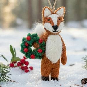 FINLEY FESTIVE FOX ORNAMENT, a handmade felt fox holding a small green wreath with red berries, standing in the snow.