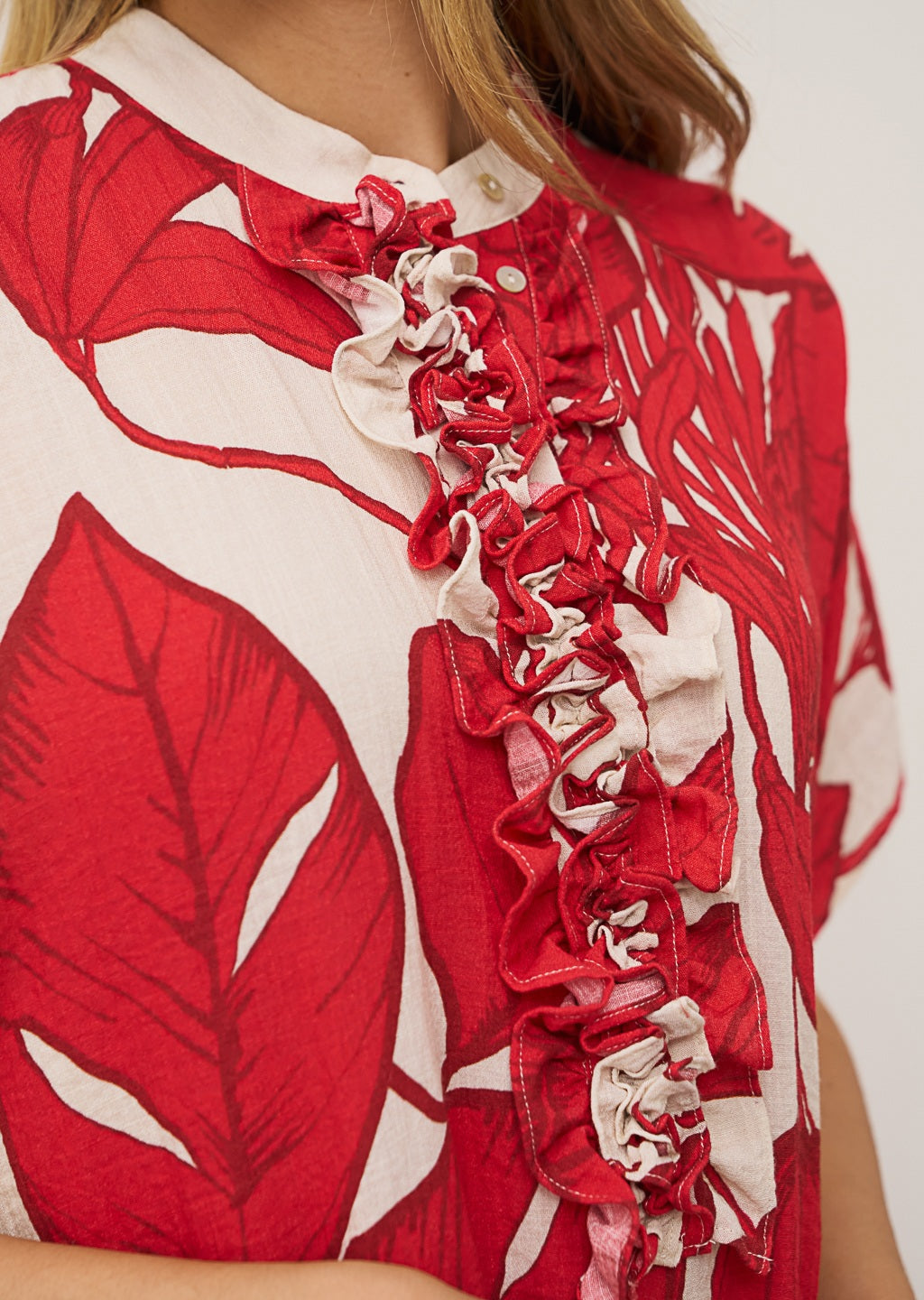 A woman wears the DAISY DRESS IN RED PRINT by 0039 ITALY—a loose, O-shape silhouette with a bold red and white leaf pattern—paired with red flip-flops, standing against a plain light wall and gazing slightly to the side.