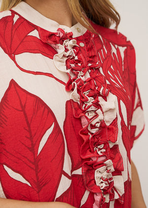 A woman wears the DAISY DRESS IN RED PRINT by 0039 ITALY—a loose, O-shape silhouette with a bold red and white leaf pattern—paired with red flip-flops, standing against a plain light wall and gazing slightly to the side.