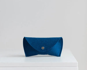 Three WOOLBERRY WOOL FELT GLASSES CASES in navy, bright, and light blue with magnetic closures are arranged on a white surface near clear sunglasses, framed by white-blue hydrangeas.