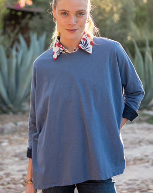 A woman stands outdoors in the MERSEA - CATALINA SLUB TEE IN MOON BLUE by MERSEA, a floral-patterned collar peeking out as she smiles softly amid sunlight and greenery.