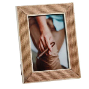 Two ZODAX photo frames—a 5x7 ribbed mango wood frame with a white bone border—rest on a beige surface. One frame displays an image of a person holding a cigar and glass; the other features a woman with long hair seated on the floor. A nearby glass filled with brown liquid completes the scene.