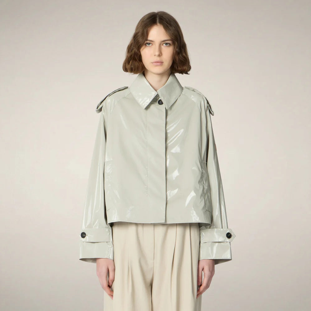 A person with chin-length brown hair wears the SAVE THE DUCK - GAIA JACKET by SAVE THE DUCK, a shiny light-gray, cropped trench-style women's jacket over a white top, paired with high-waisted beige trousers and beige shoes against a plain background.