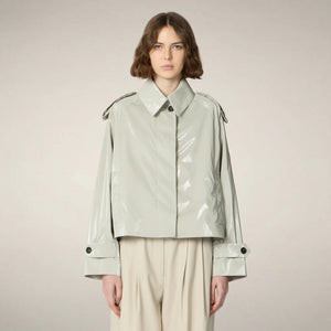 A person with chin-length brown hair wears the SAVE THE DUCK - GAIA JACKET by SAVE THE DUCK, a shiny light-gray, cropped trench-style women's jacket over a white top, paired with high-waisted beige trousers and beige shoes against a plain background.