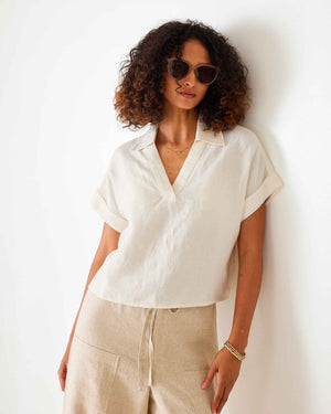 A woman with curly hair wears sunglasses and the MERSEA HADLEY BOXY LINEN SHIRT in cream, featuring loose short sleeves, paired with beige high-waisted pants with front pockets, standing against a white background.