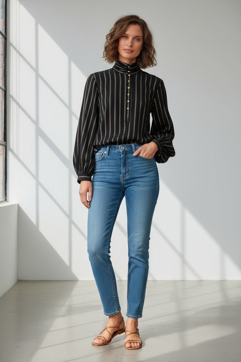 A woman with wavy brown hair, arms crossed, wears the MOMONI Thierry Blouse in lurex stripe gauze and a patterned skirt, standing against a pale background.
